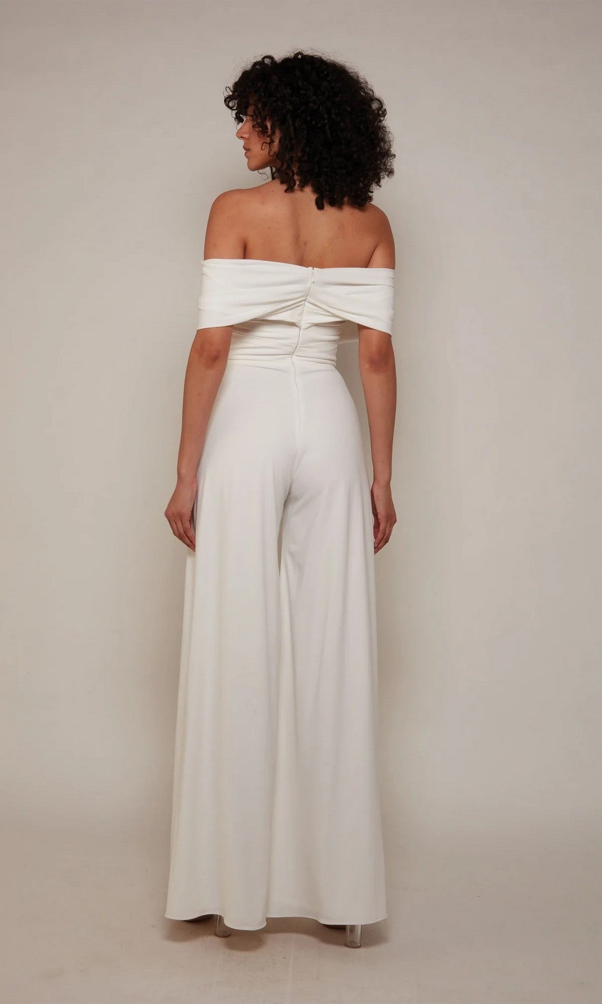 A person with curly hair stands facing away, wearing the Alyce 70051 long white off-the-shoulder satin jumpsuit with wide-leg pants against a plain light background.